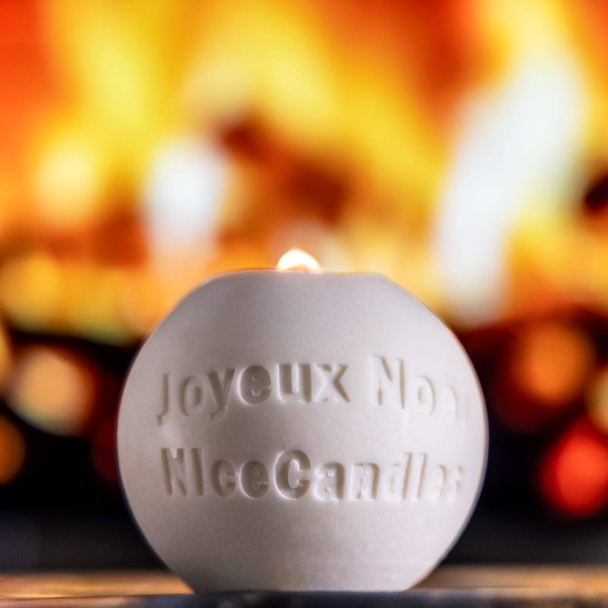 Bougie artisanale en forme de boule de Noël avec inscriptions 'Joyeux Noël' et 'NiceCandles', allumée devant un fond chaleureux de feu de cheminée, idéale pour créer une ambiance festive et conviviale.