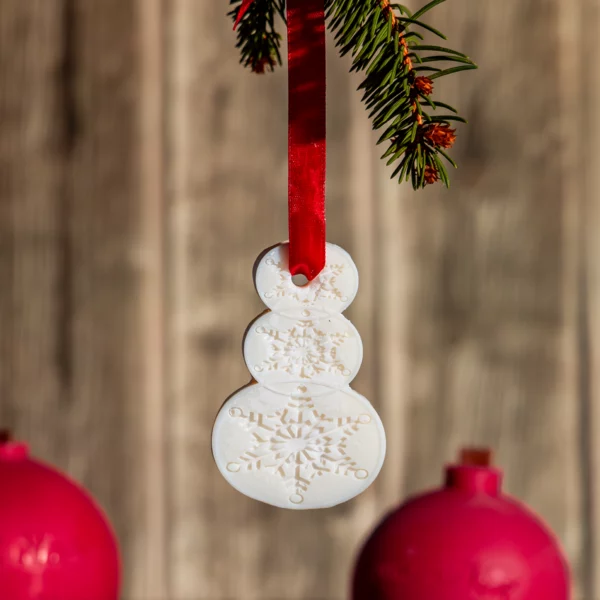 Suspension parfumée en forme de bonhomme de neige en cire blanche avec motifs de flocons de neige, suspendue par un ruban rouge. Idéale comme décoration de Noël parfumée pour sapins, armoires et petits espaces.